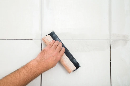 Une main masculine applique du joint silicone blanc sur carrelage mural avec une spatule &agrave; jointoyer